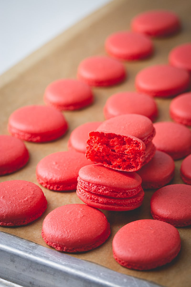 Red macaron shells on a baking sheet lined with parchment paper with one shell halved.