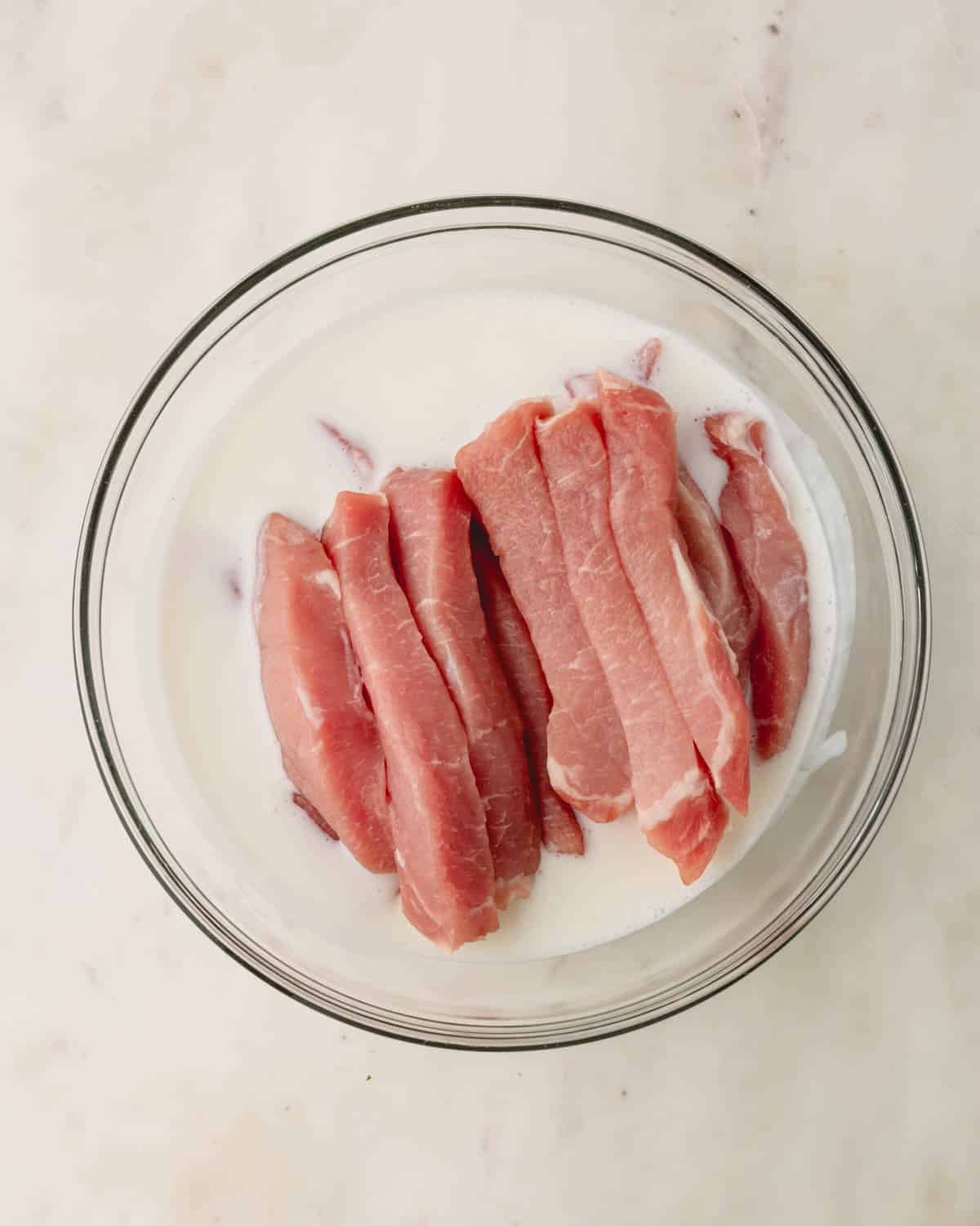 Raw strips of pork tenderloin in a bowl of buttermilk.