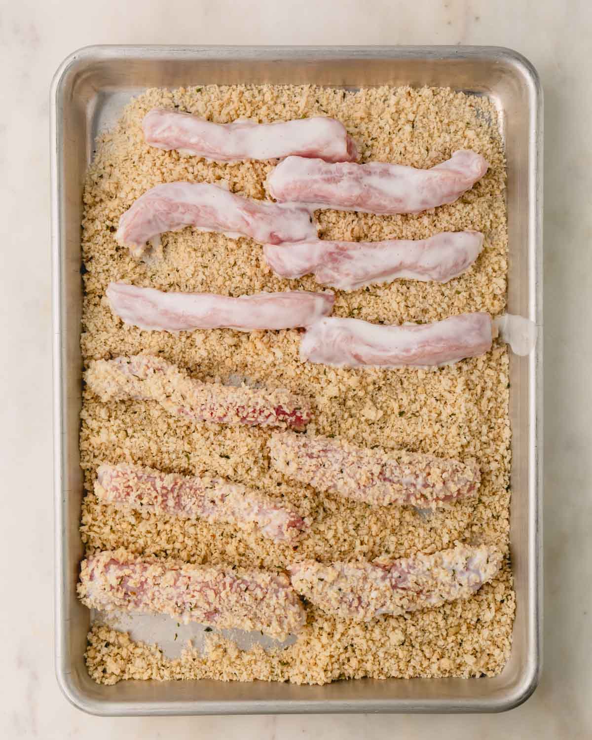 Buttermilk coated pork strips on a baking dish with breadcrumbs.