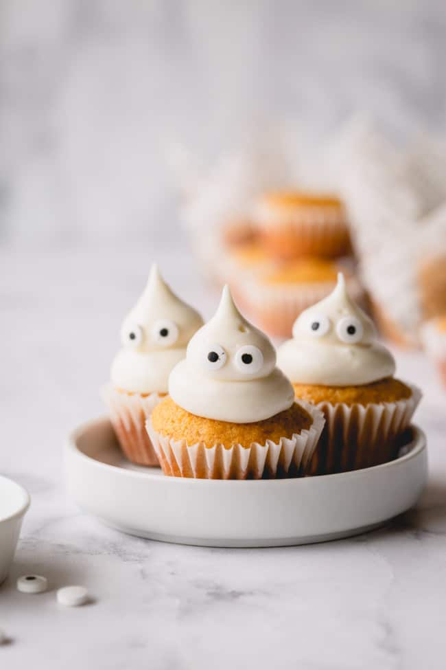 3 mini pumpkin muffins on a white plate topped with domed cream cheese frosting and candy eyes.