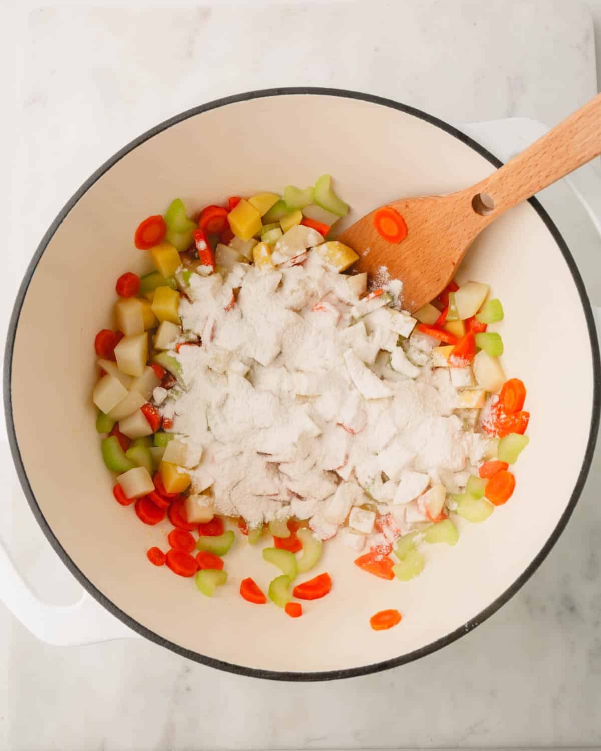 Chopped carrots, celery, and potatoes are topped with flour in a Dutch oven with a wooden spoon.