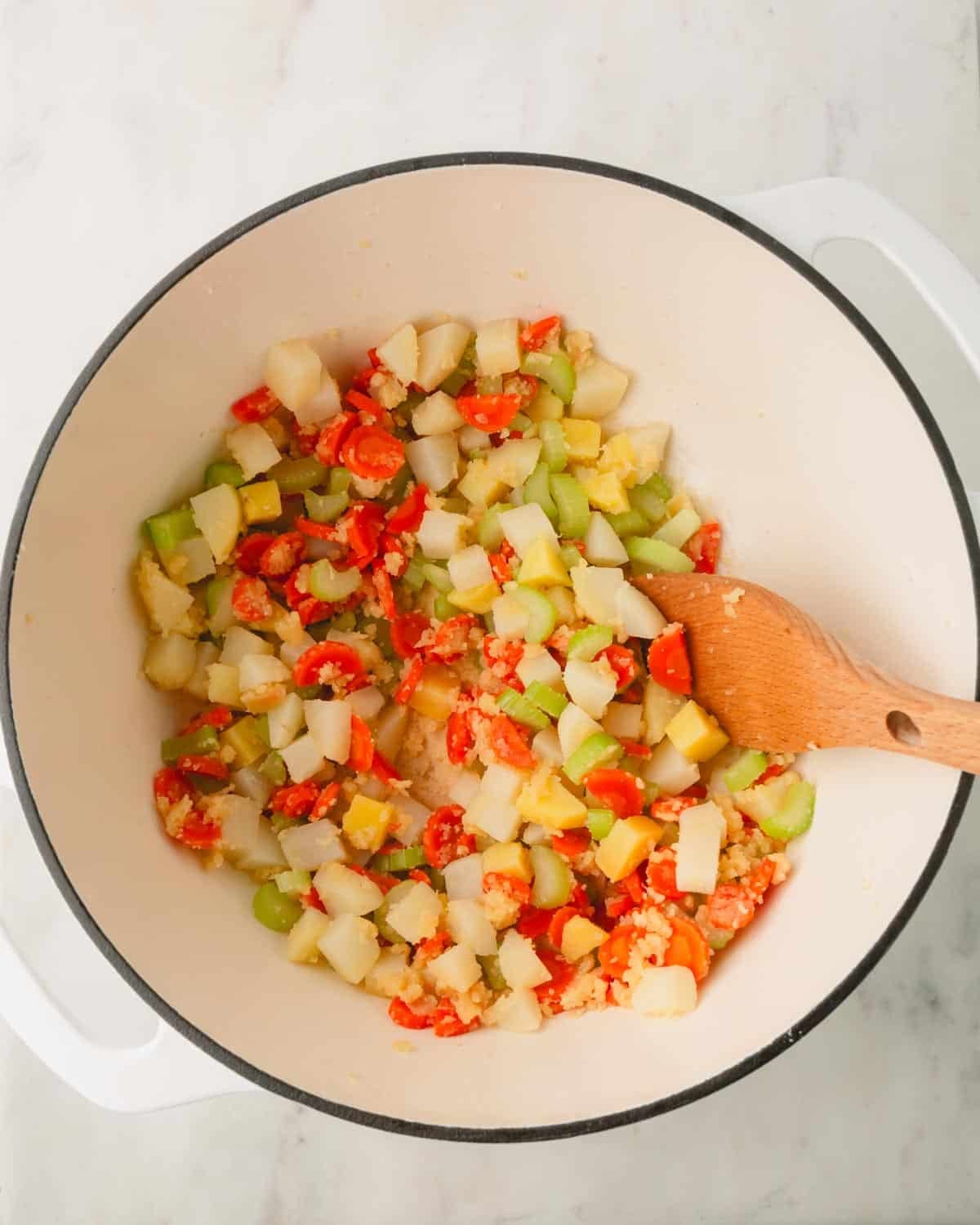 Chopped carrots, celery, and potatoes are coated with flour in a Dutch oven with a wooden spoon.