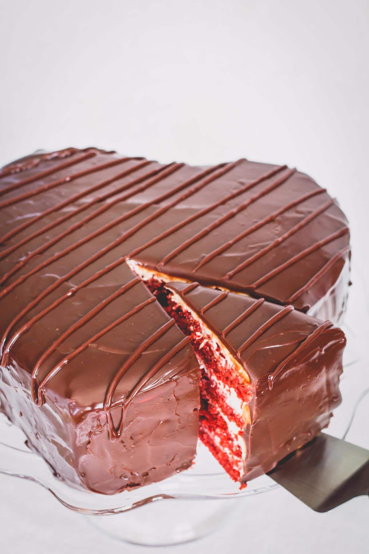 Heart shaped chocolate covered red velvet cake on a glass platter with a slice cut.