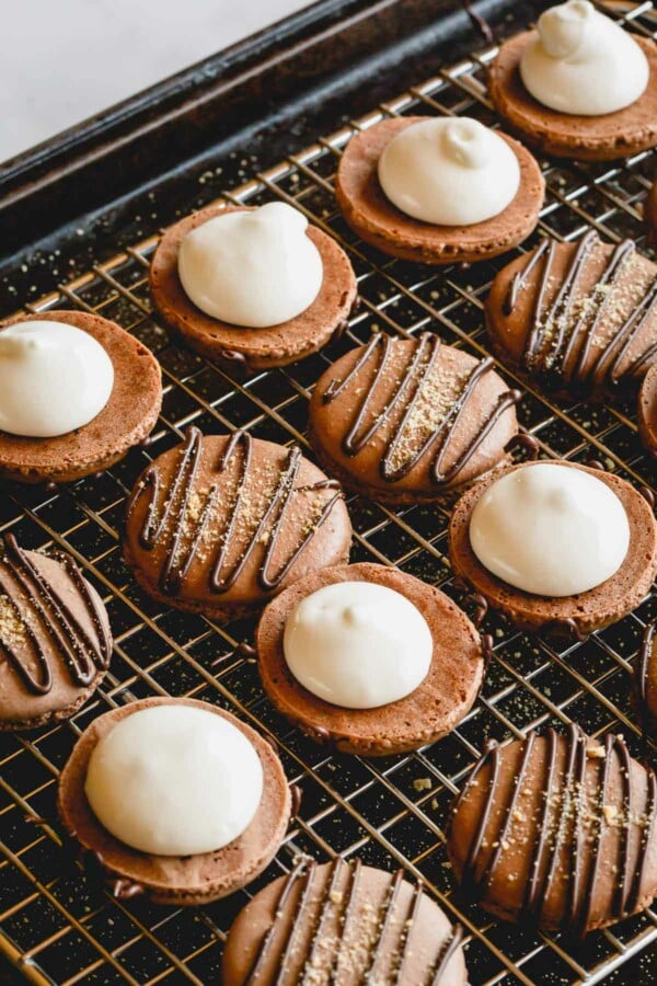 Chocolate macaron shells on a wire rack with a dollop of marshmallow filling.