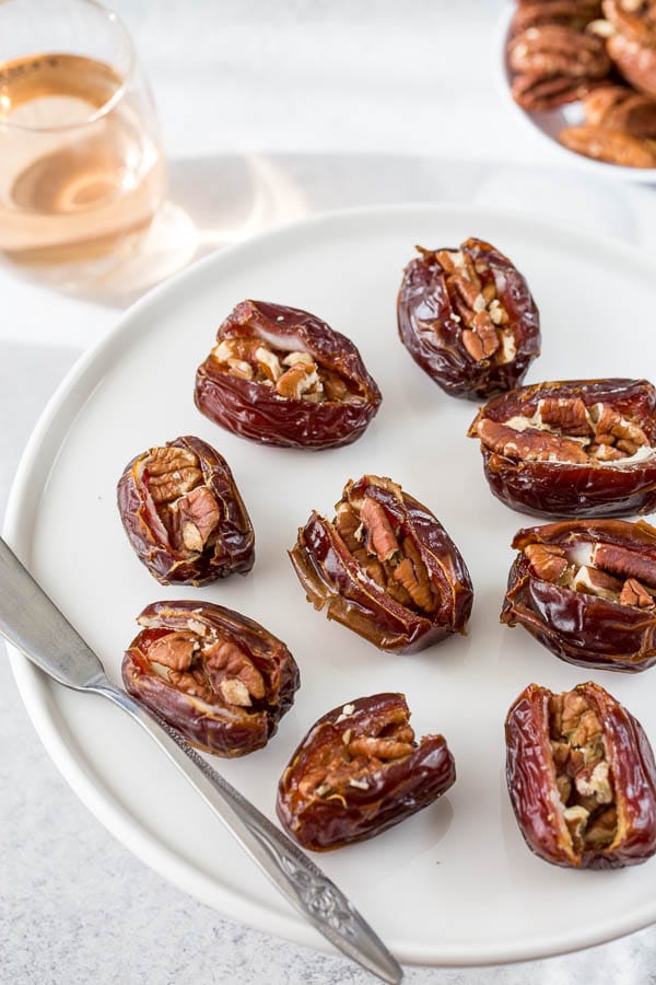 Roasted medjool dates stuffed with cream cheese and pecans arranged on a platter with a glass of wine. Roasted stuffed dates are addicting bites of all time!!! Roasting turns sweet medjool dates into gooey "caramels" that pairs beautifully with toasted nuts inside. #datesnacks #dateappetizer #easyappetizer