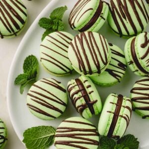 Green mint macarons filled with dark mint ganache and drizzled with chocolate arranged on a white oval plate and garnished with mint leaves.