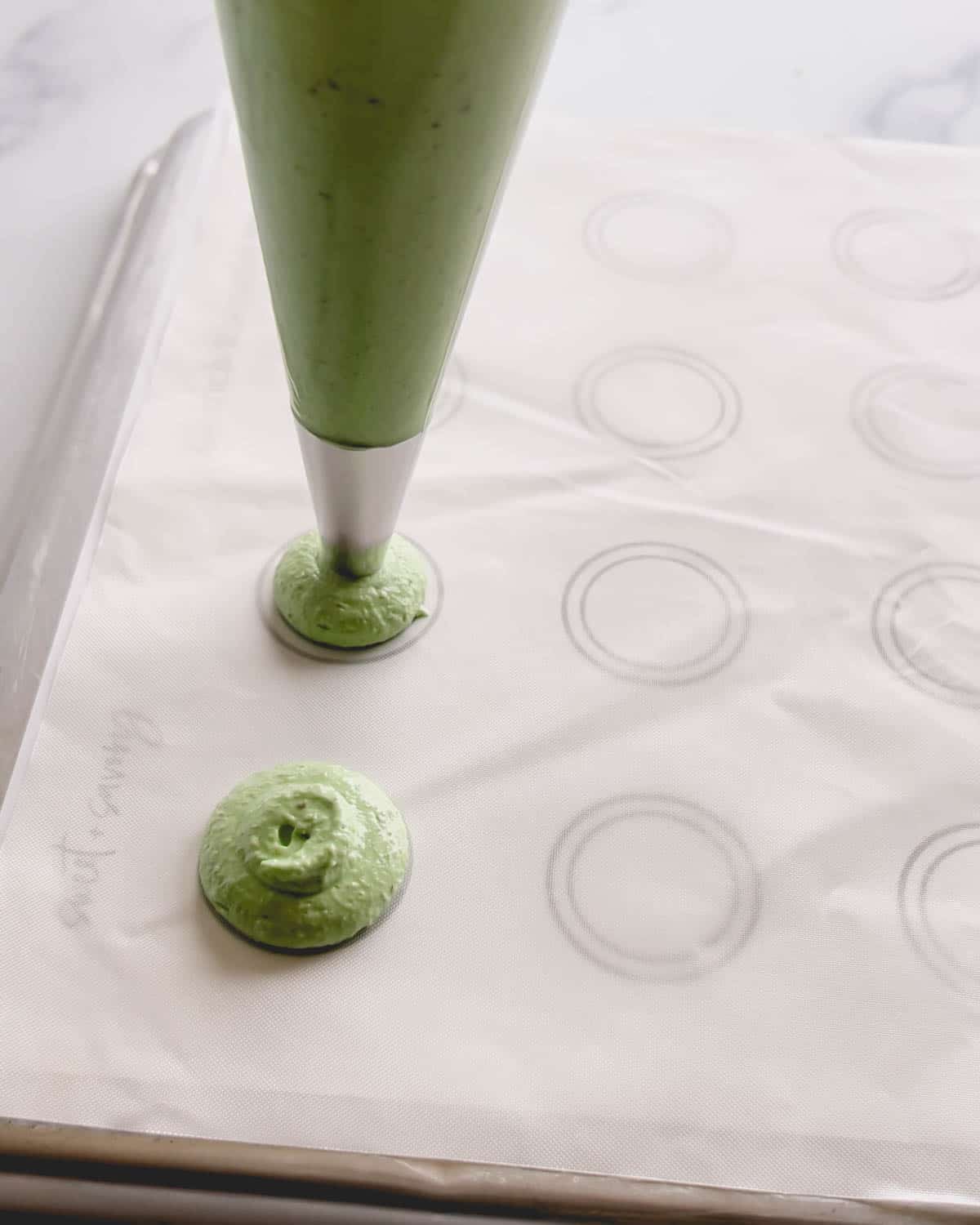 Green macaron shells being piped on a white surface with circle pattern.
