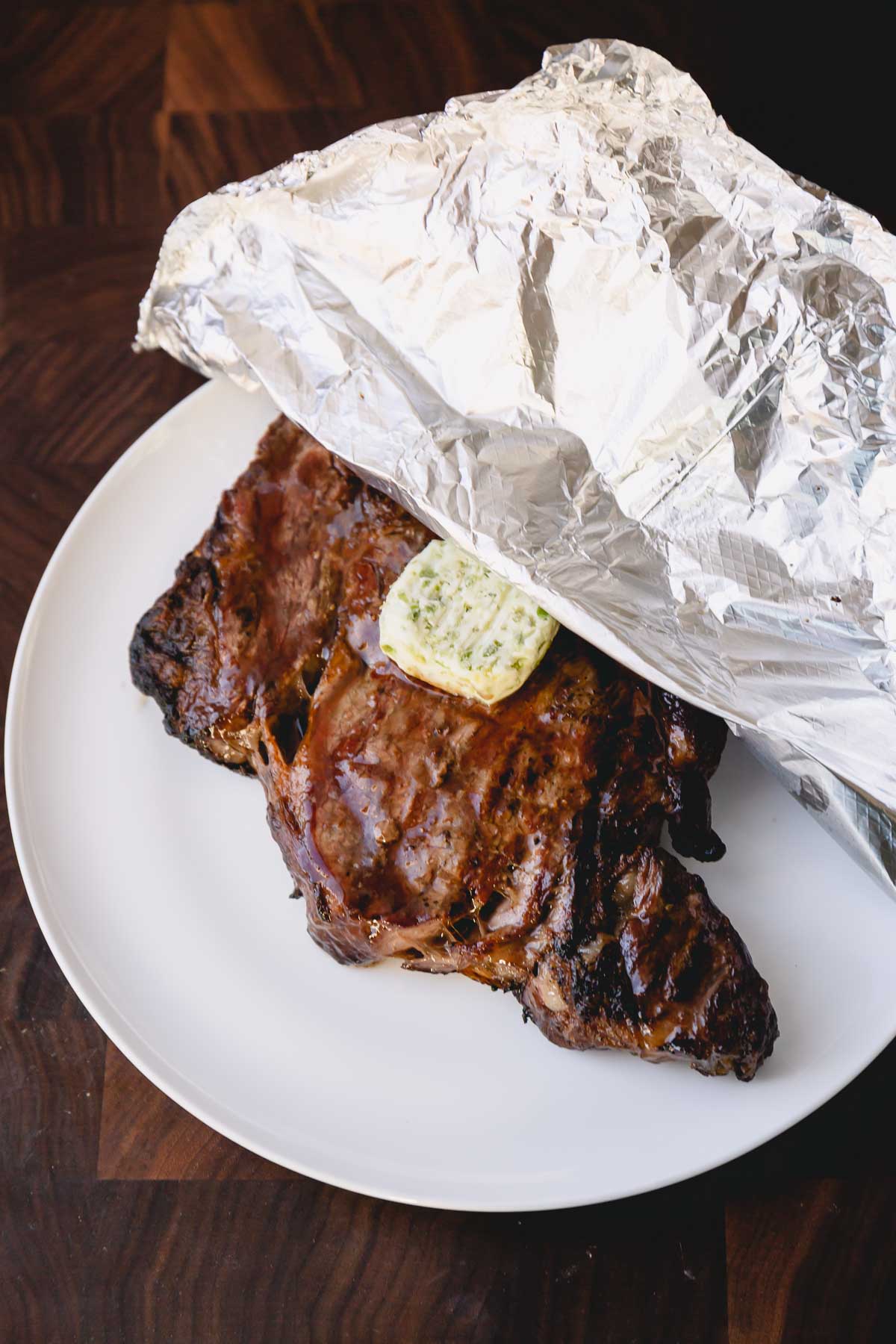 Grilled ribeye steak with a slice of compound butter on a plate half covered with foil