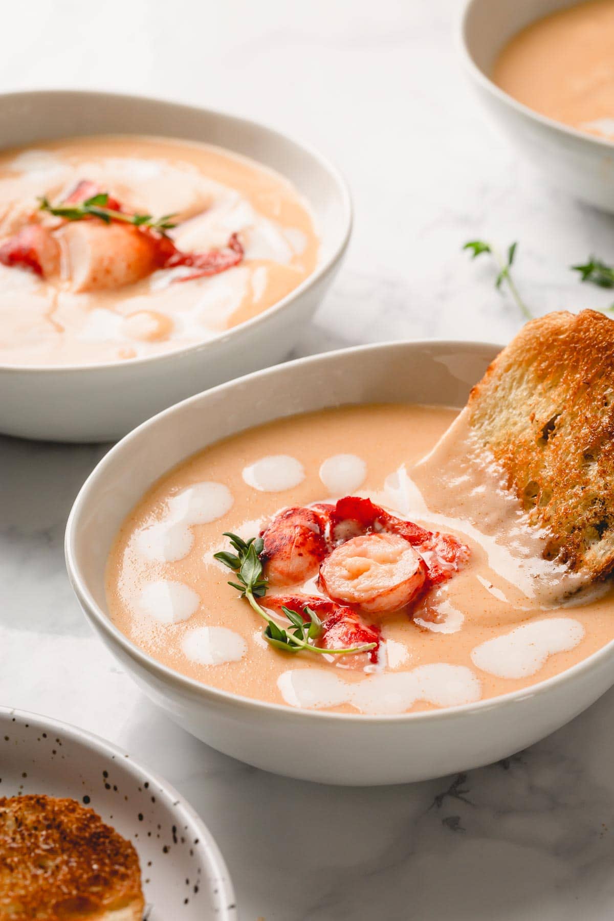 A bowl of lobster bisque garnished with heavy cream, lobster chunks, fresh thyme sprig and a side of toasted bread.