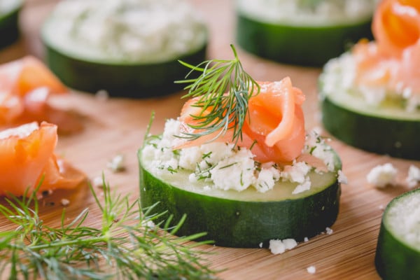 A cucumber slice topped with feta cheese, dill and salmon on a wooden serving platter.