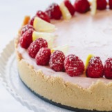 a whole lemon raspberry cheesecake topped with fresh raspberries on a white serving platter.