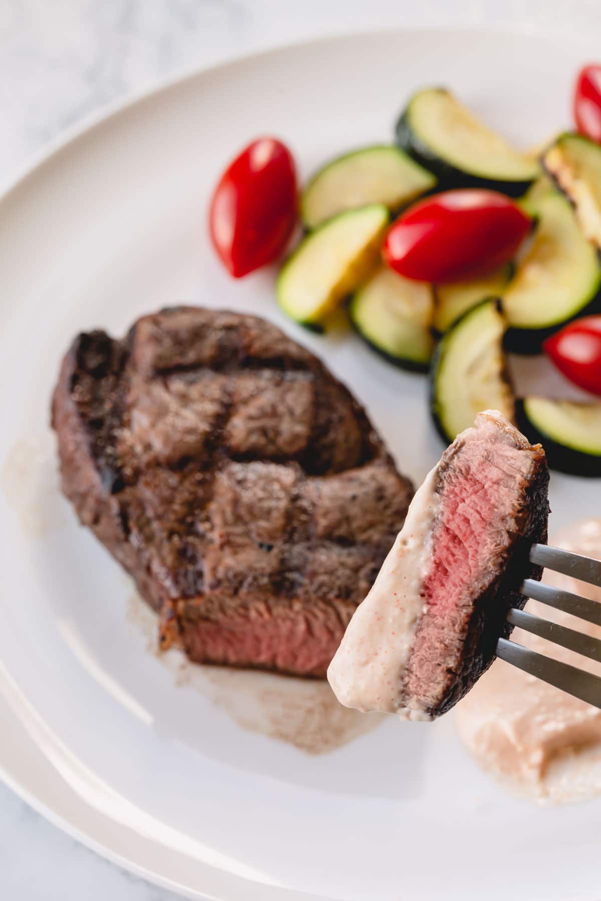 A whole grilled filet mignon on a white plate with zucchini and cherry tomatoes on the side.