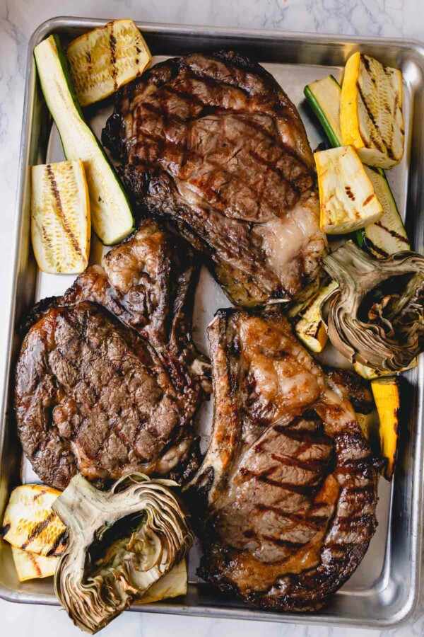 3 grilled ribeye steaks on a baking sheet with grilled zucchini and artichokes.
