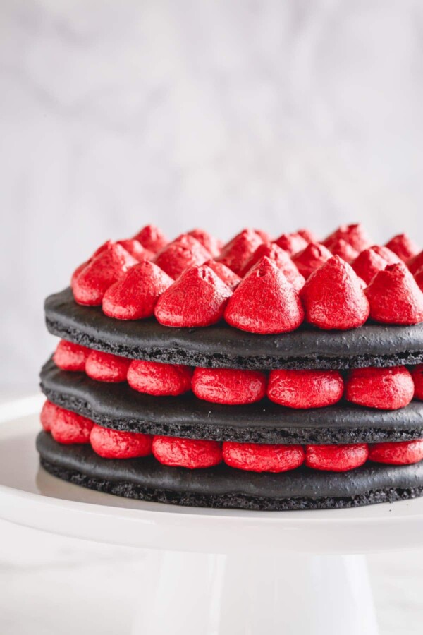 A 3-layer black macaron cake filled with red cream cheese filling on a white cake stand.