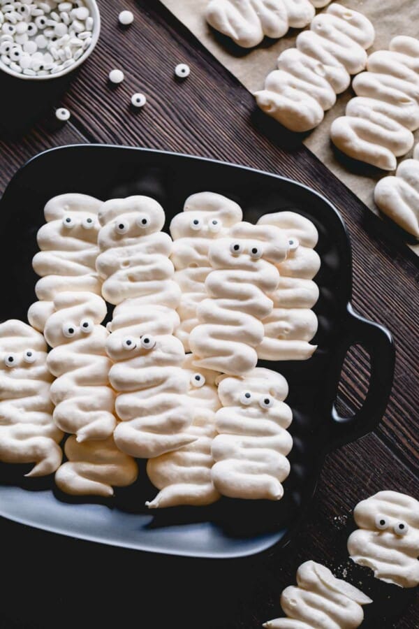 Mummy meringue cookies with googly eyes on a black plate.