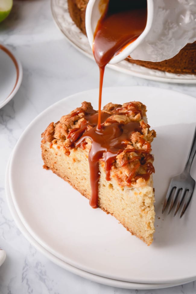 A slice of apple coffee cake on a white plate with caramel sauce being pour over it.