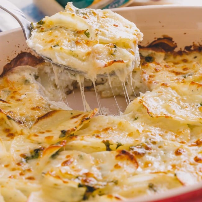 Cheesy scalloped potatoes in a casserole dish with a serving spoon taking a piece.