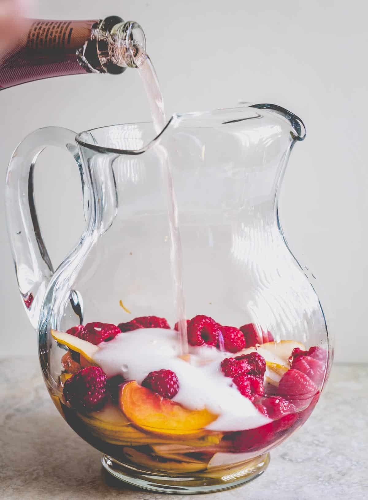 A glass pitcher filled with peach slices and raspberries and rose wine pouring in.
