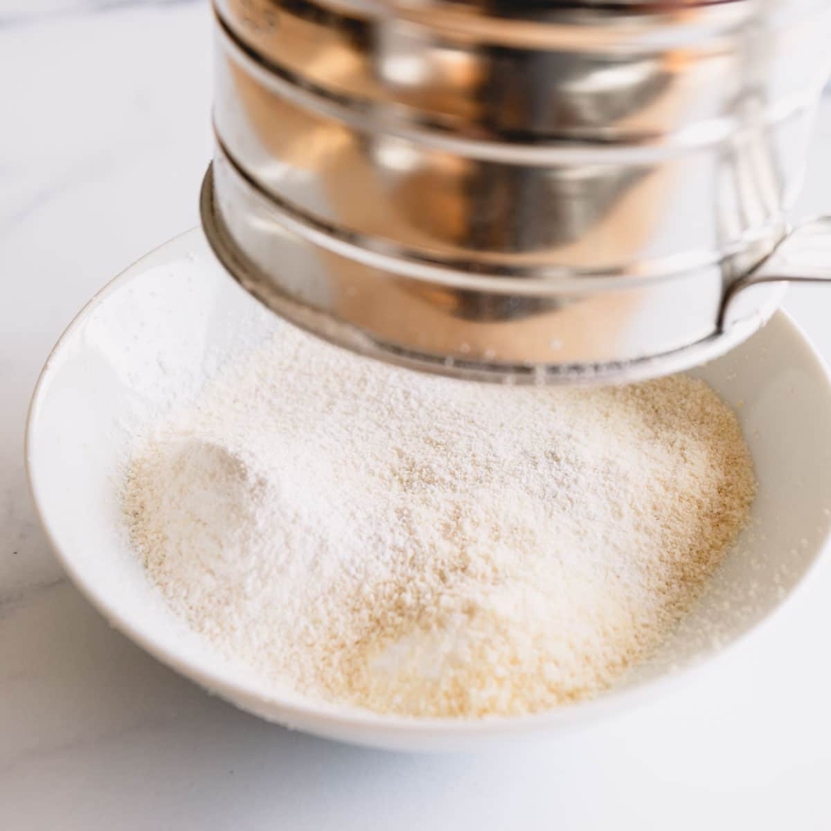 Almond flour and powdered sugar being sifted into a white bowl.