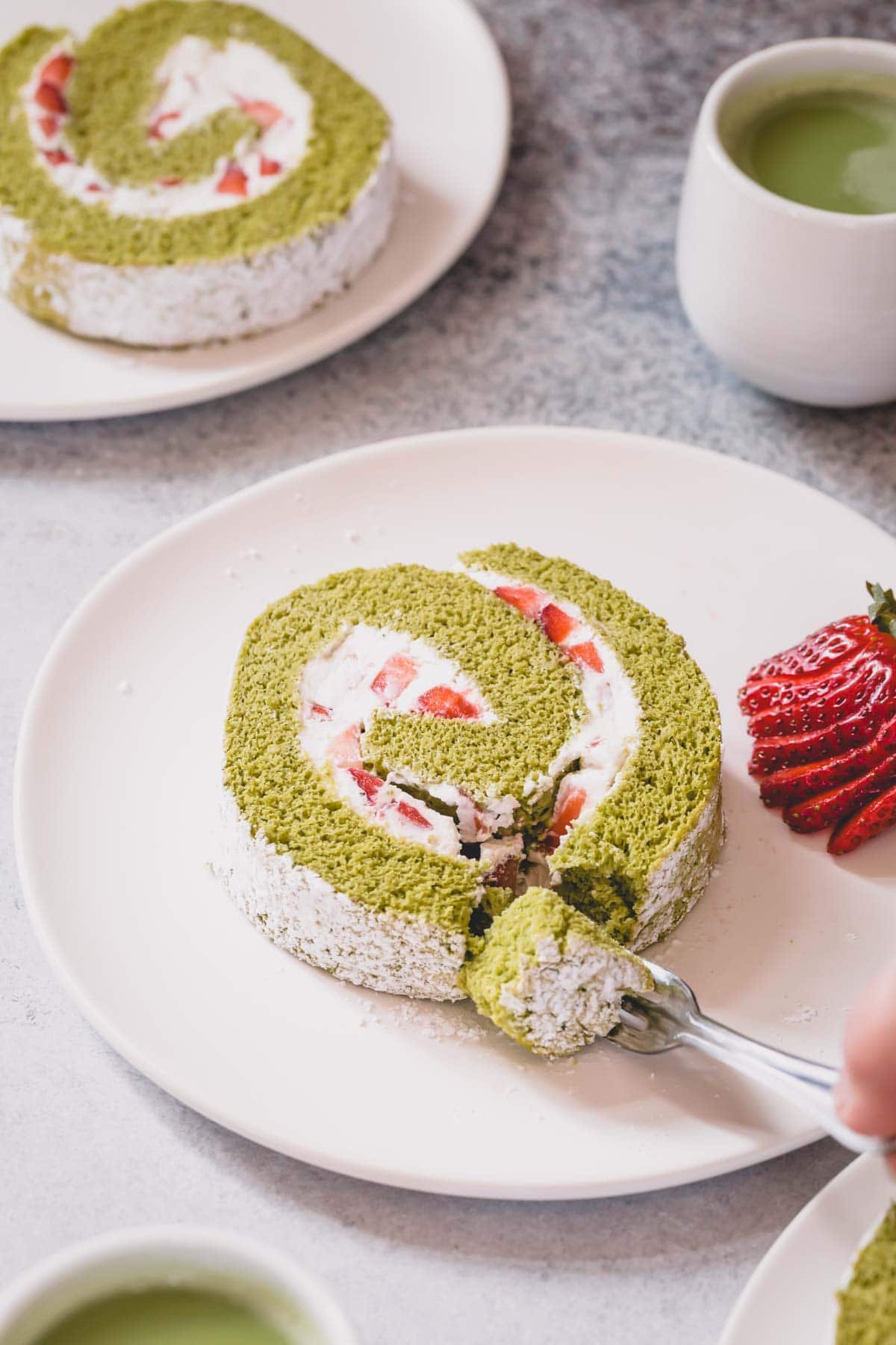 A slice of strawberry matcha roll cake on a white serving plate with a a little slice cut with a fork.