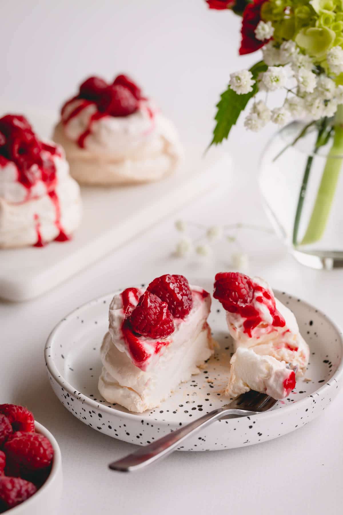 Mini pavlova, topped with raspberry whipped cream, raspberry sauce and fresh raspberries, sliced in half.