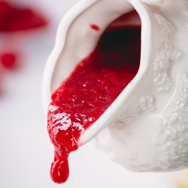 Raspberry sauce is being poured from a white sauce pitcher.