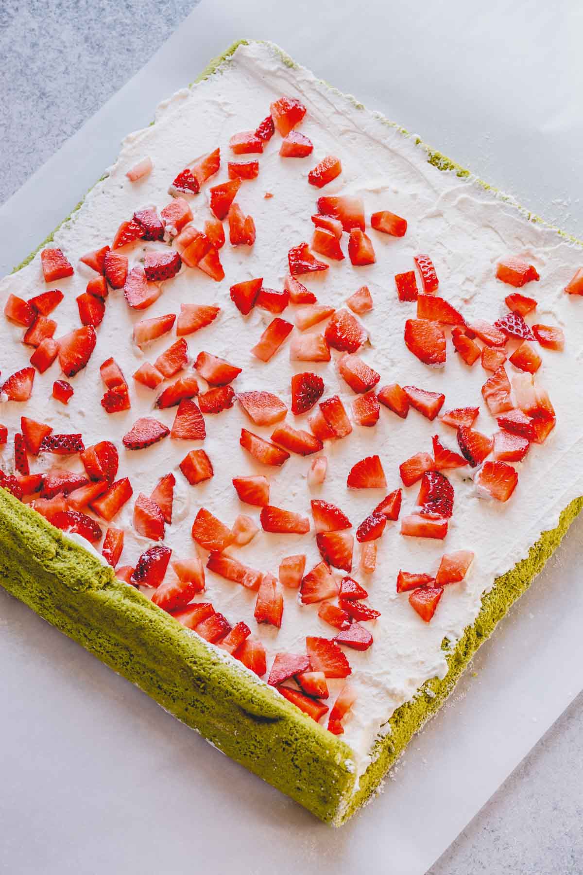 Sheet of green sponge cake with whipped cream spread all over and diced strawberries scattered over the cream.