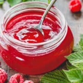 A glass jar of raspberry jelly with a spoon dip in it.