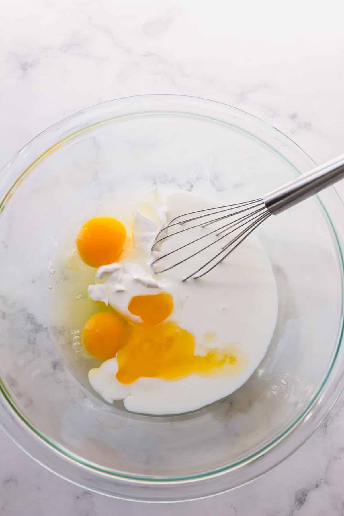 Eggs, buttermilk, and sour cream in a glass bowl with a whisk.