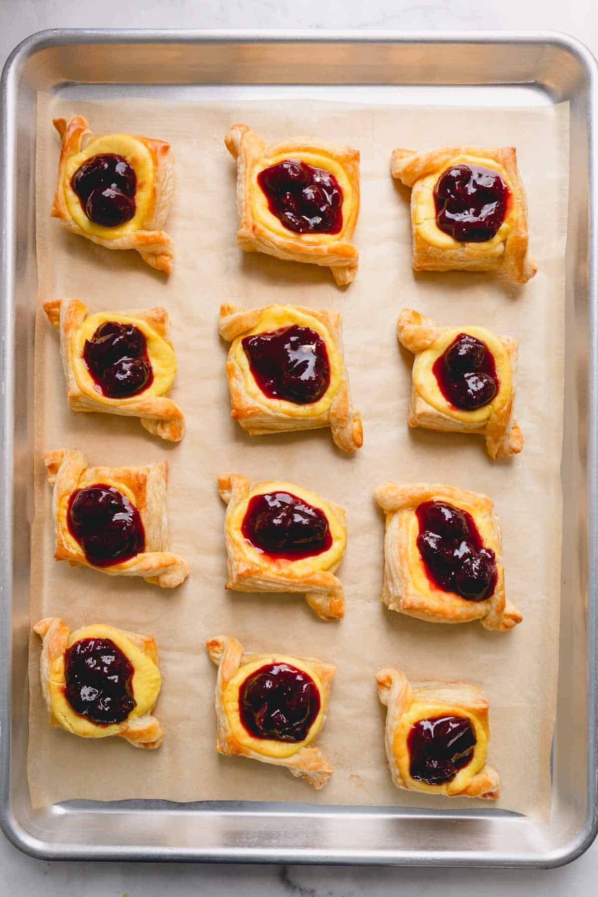 A parchment paper-lined baking sheet with 12 cherry cheese danishes.