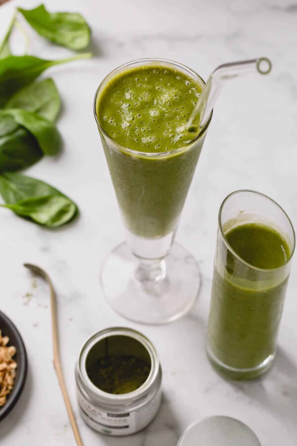 Two glasses full of a coconut green smoothie with an open jar of matcha powder.