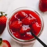Overhead image of a spoon sticking out of a glass jar full of chunky strawberry sauce.