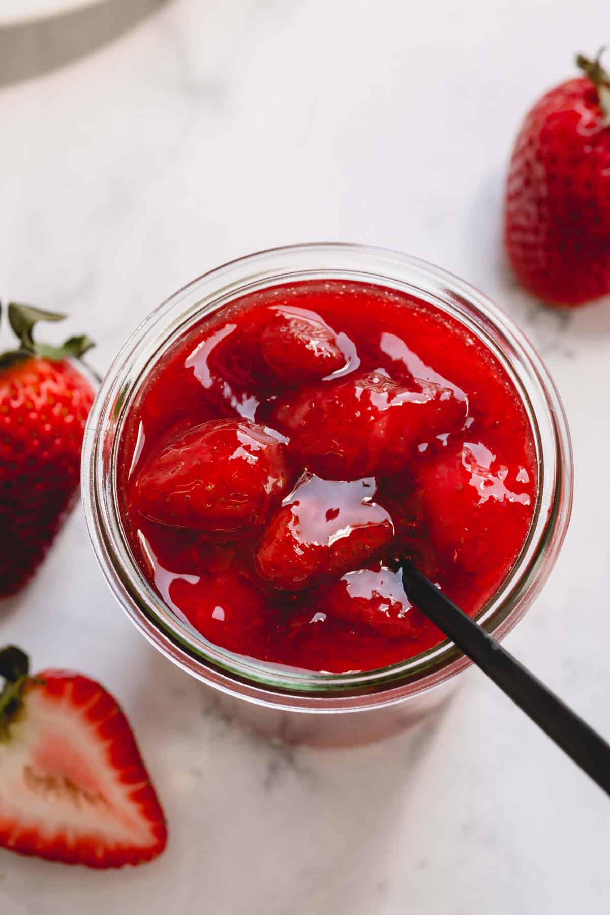 Overhead image of a spoon sticking out of a glass jar full of chunky strawberry sauce.