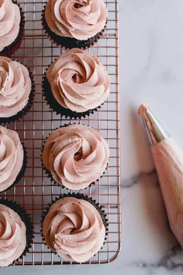 Chocolate cupcakes on a wire rack topped with strawberry whipped cream frosting.