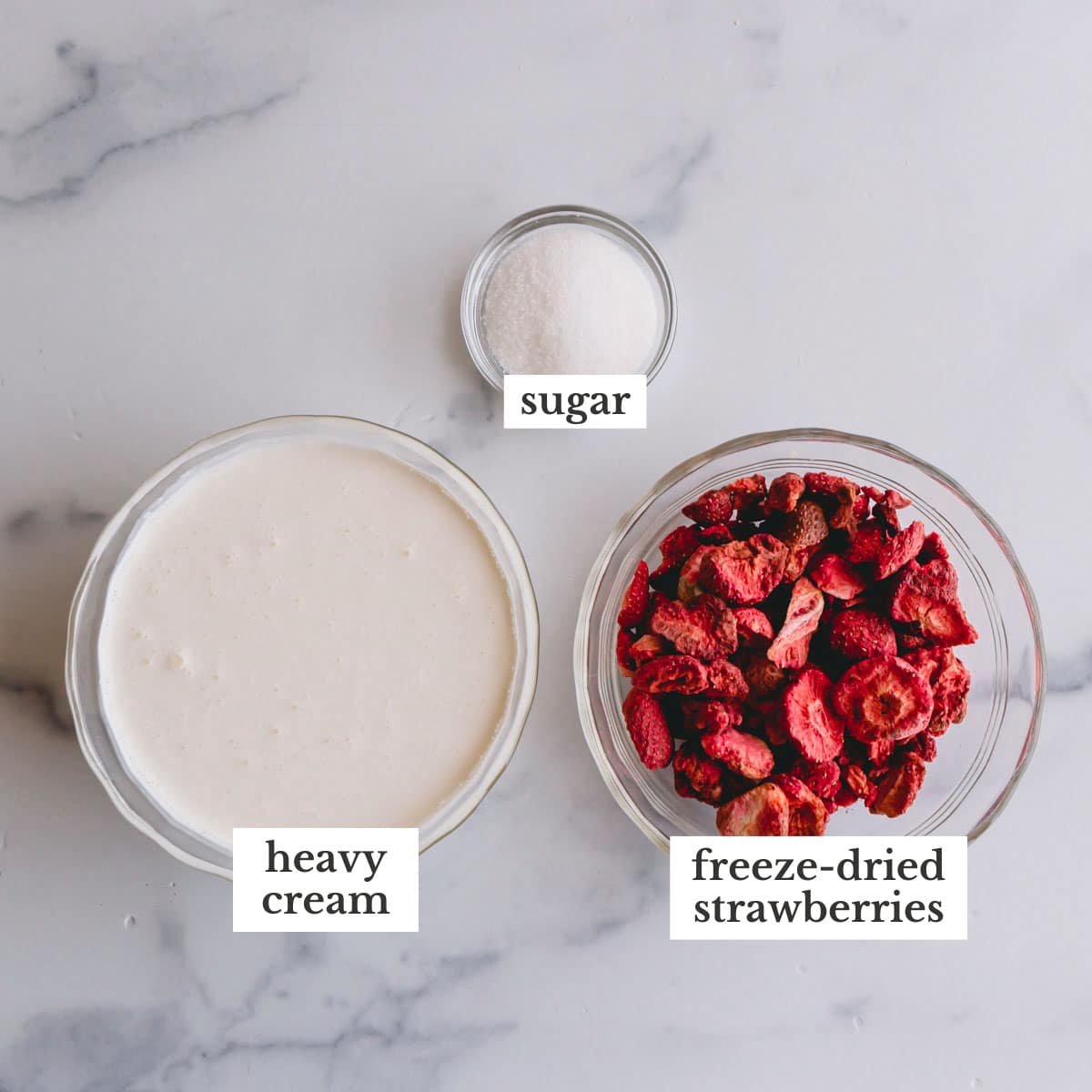 Overhead image of a bowl of heavy cream, a small bowl of sugar, and a bowl of freeze-dried strawberries.