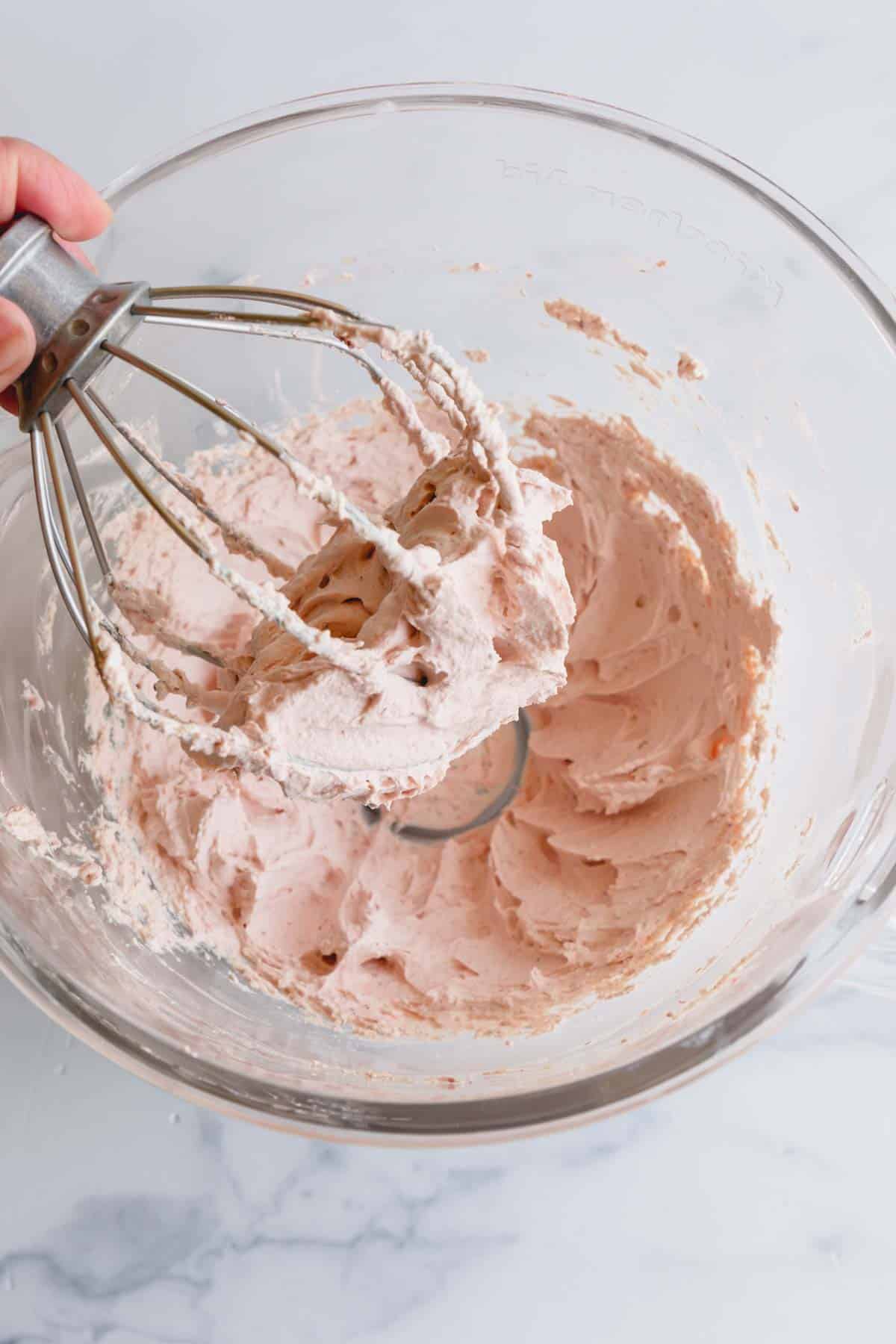 A whisk over a bowl of strawberry whipped cream frosting.