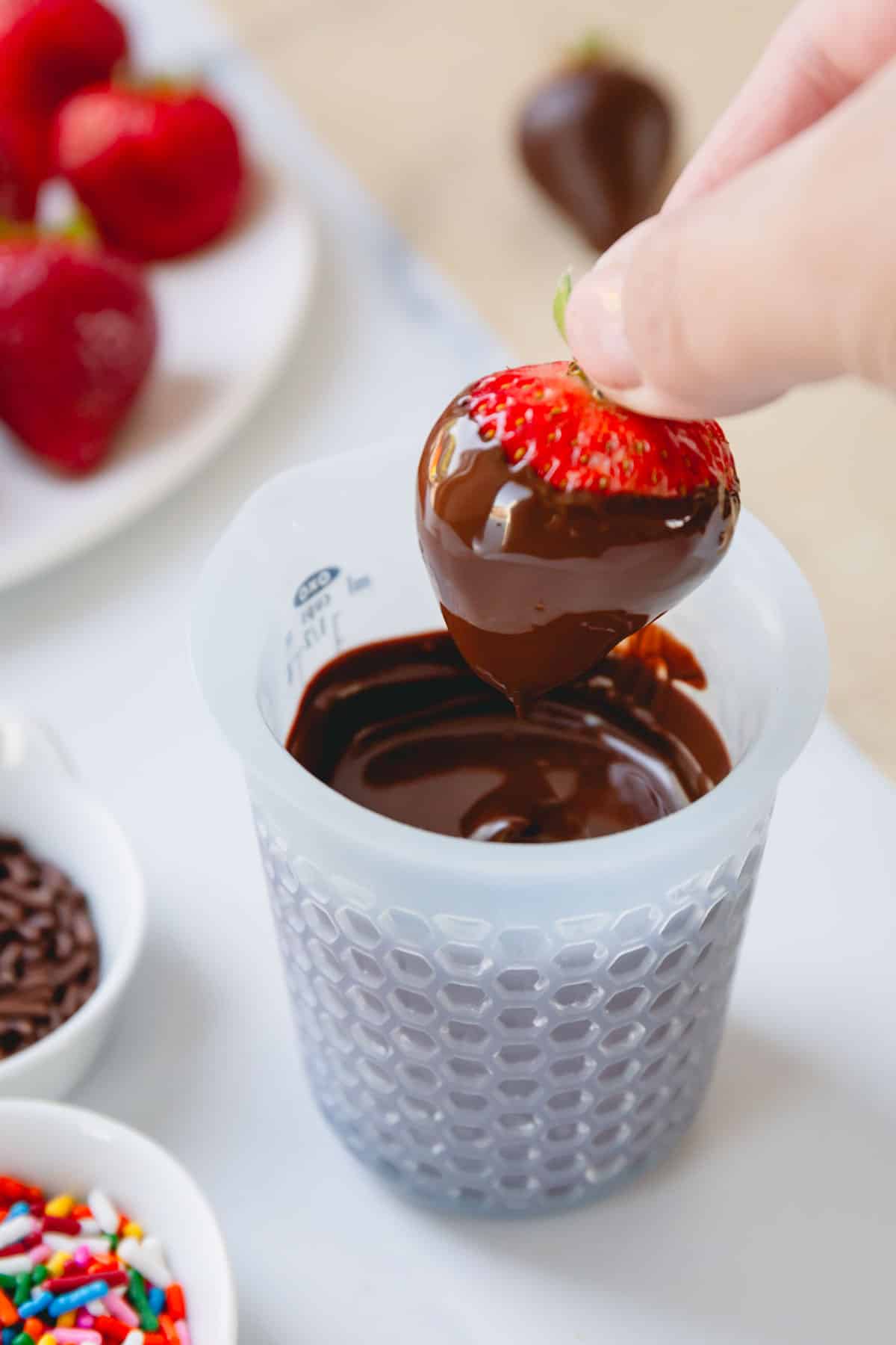 A hand holding a chocolate covered strawberry by the stem over a cup of melted chocolate.