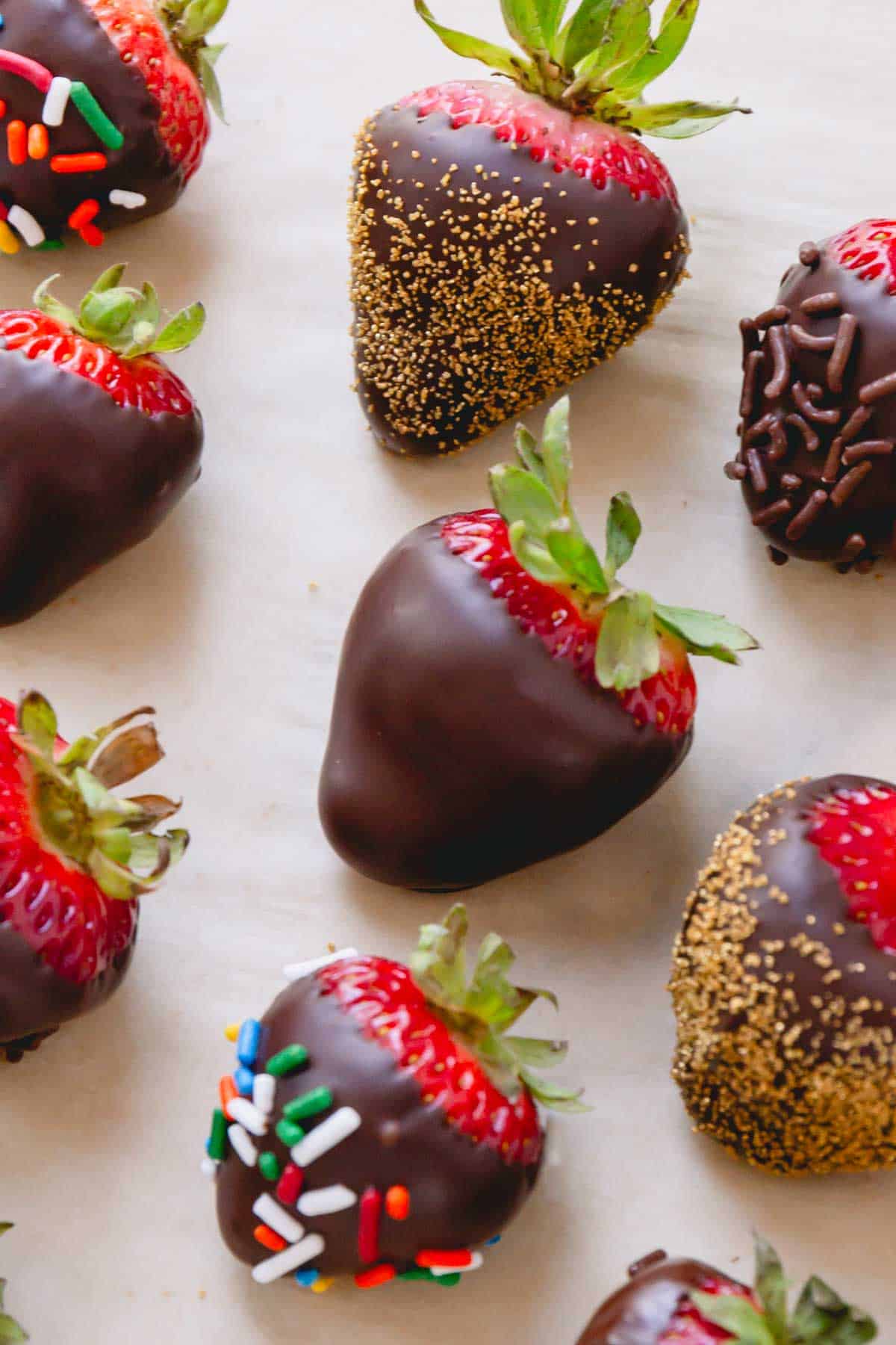 One plain chocolate covered strawberry and others with sprinkles on parchment paper.