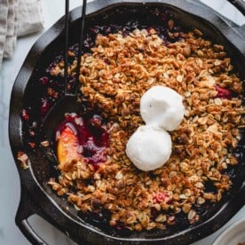 A skillet full of fruit crisp topped with two scoops of vanilla ice cream.