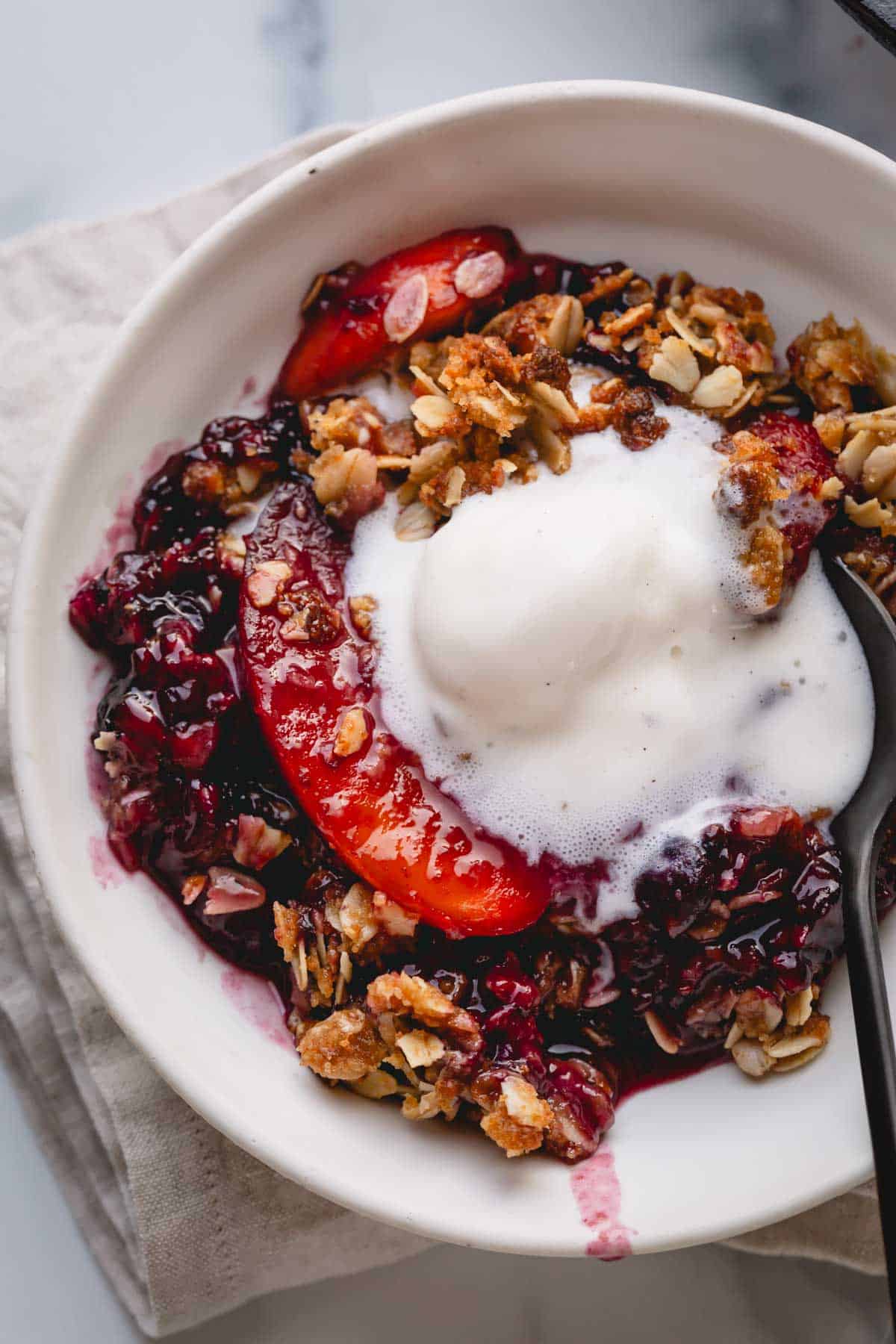 A bowl of mixed fruit crisp topped with vanilla ice cream with a spoon digging into it.