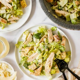 Plates of chicken Caesar salad with a bowl of salad and fresh ingredients on the side.