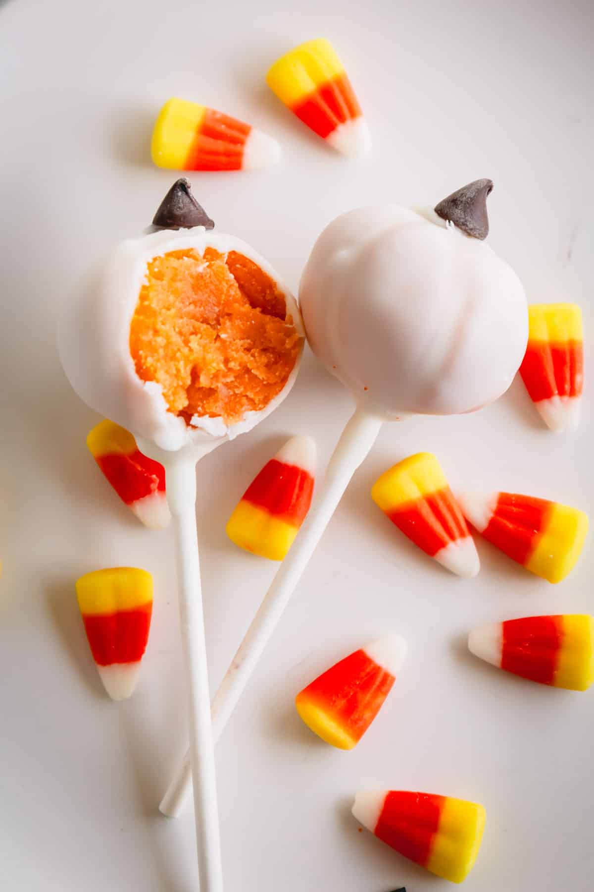 Two white pumpkin cake pops with a bite taken out of one and candy corn around them.