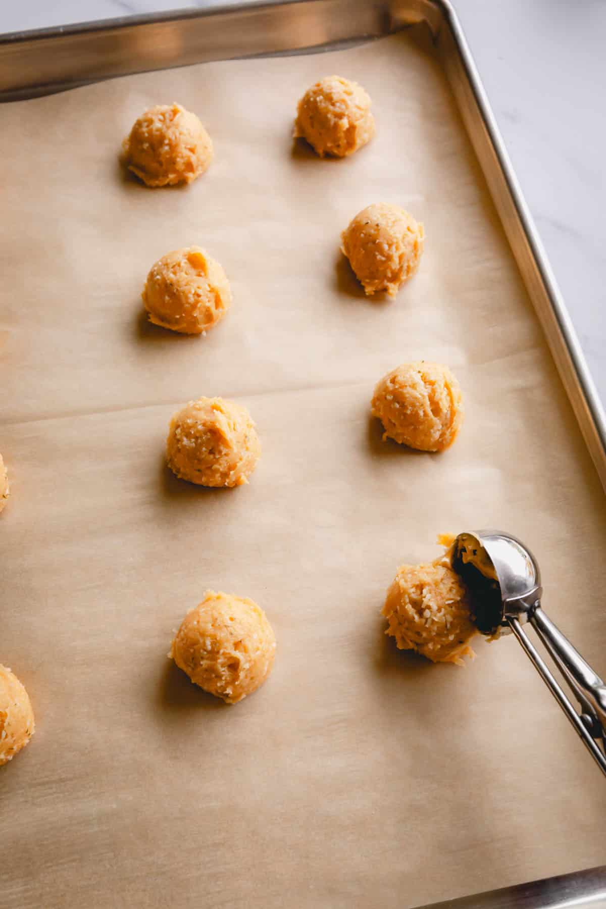 Scooping cheese puffs dough with a cookie dough scoop onto a baking sheet lined with parchment paper.