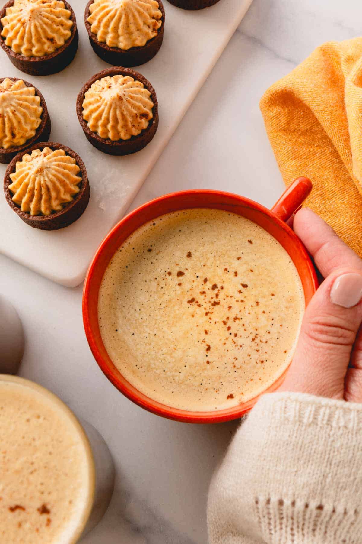 A hand holding a mug of pumpkin coffee next to chocolate cups filled with pumpkin mousse.