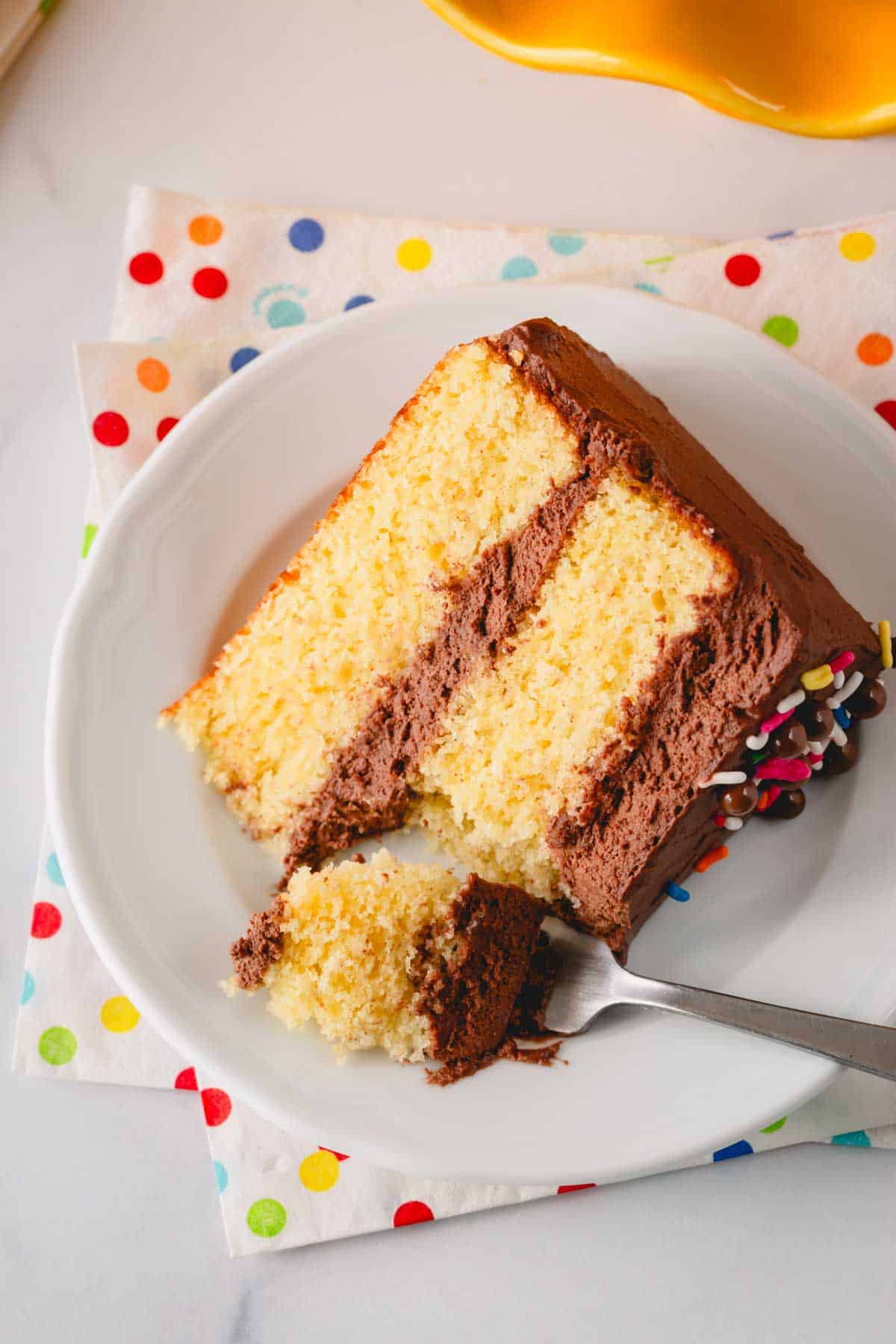 A slice of a two layer yellow cake with chocolate frosting on a plate with a fork holding a bite.