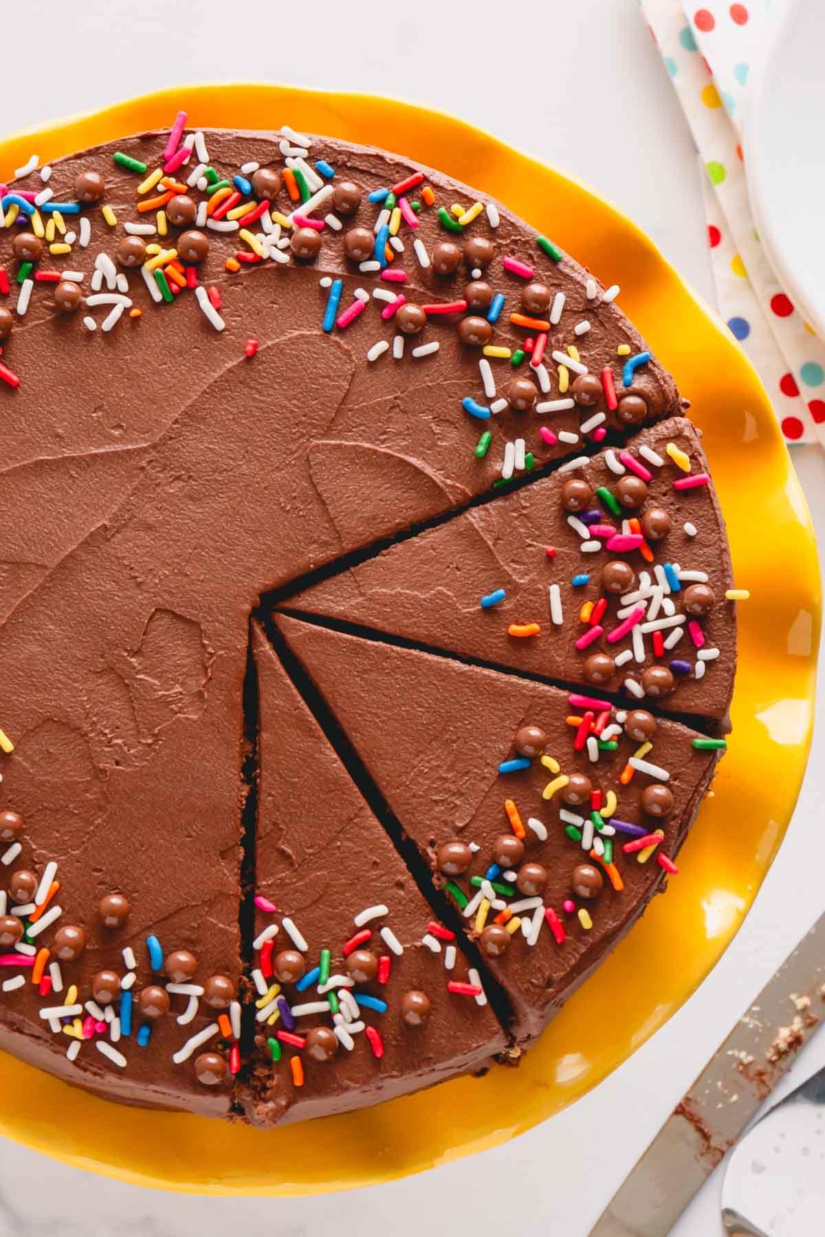 Overhead image of a chocolate frosted cake topped with sprinkles with three pieces sliced.