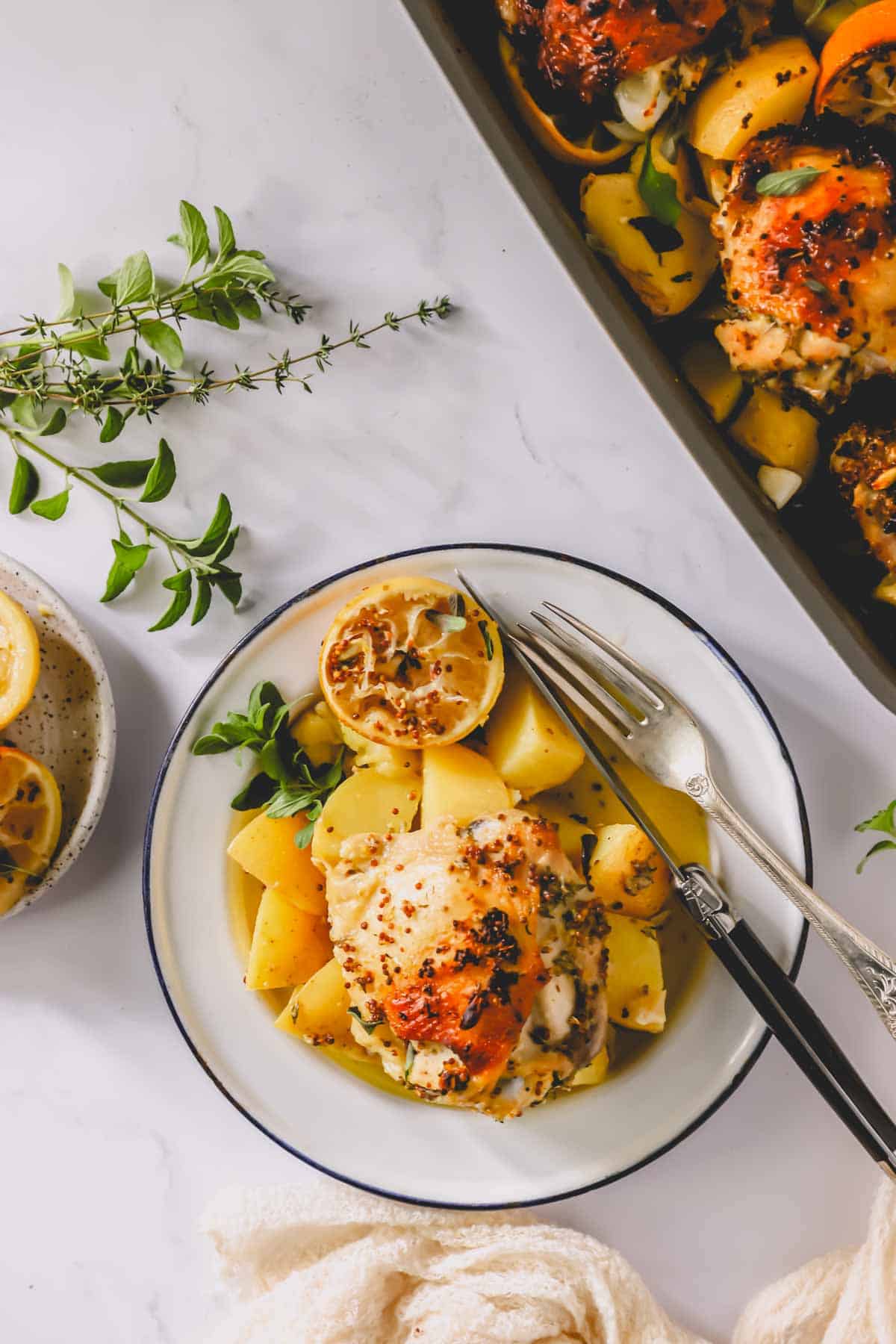 A plate with Greek lemon chicken and potatoes next to a baking dish full of the meal.