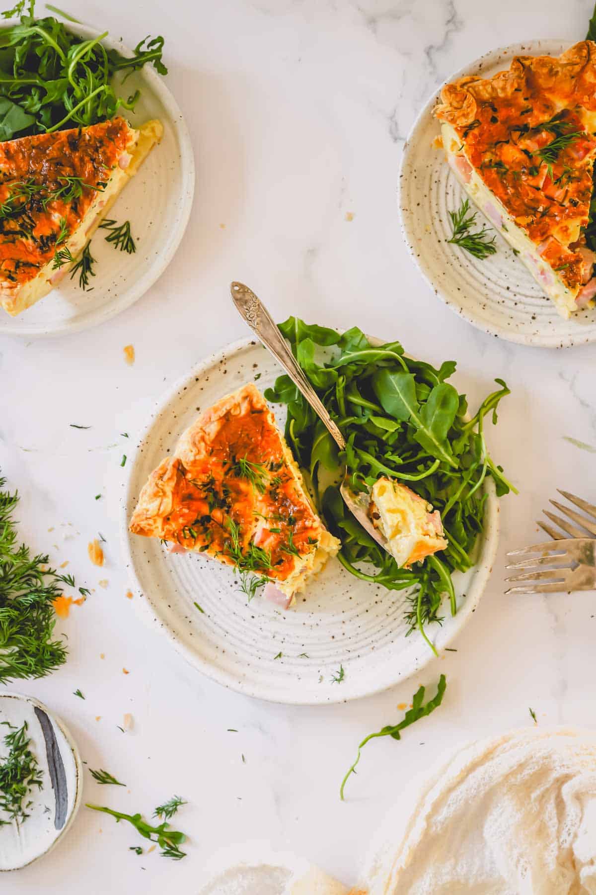 Plates with a slice of ham and cheese quiche with a aside of arugula.