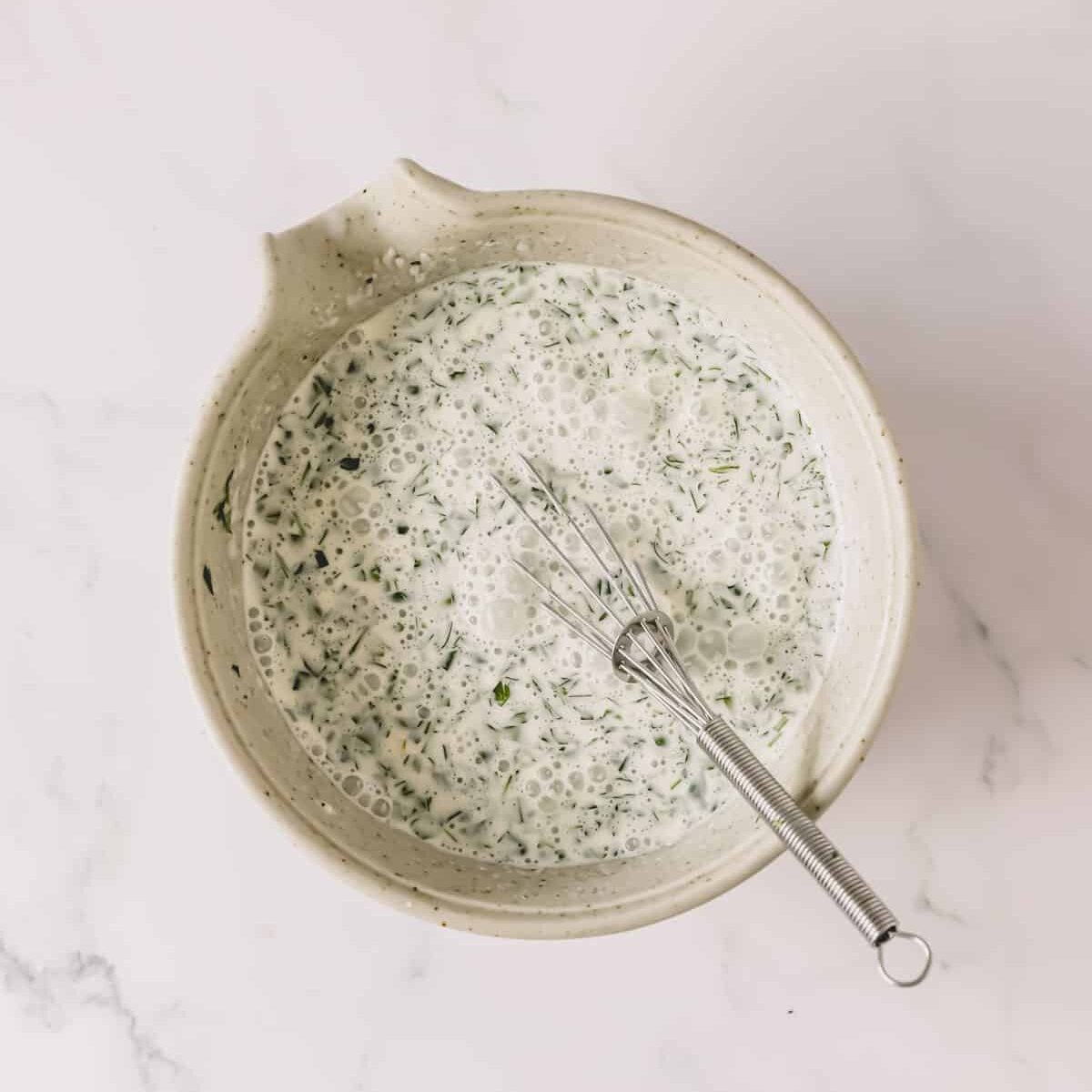 Cream and herb mixture in a bowl with a whisk.