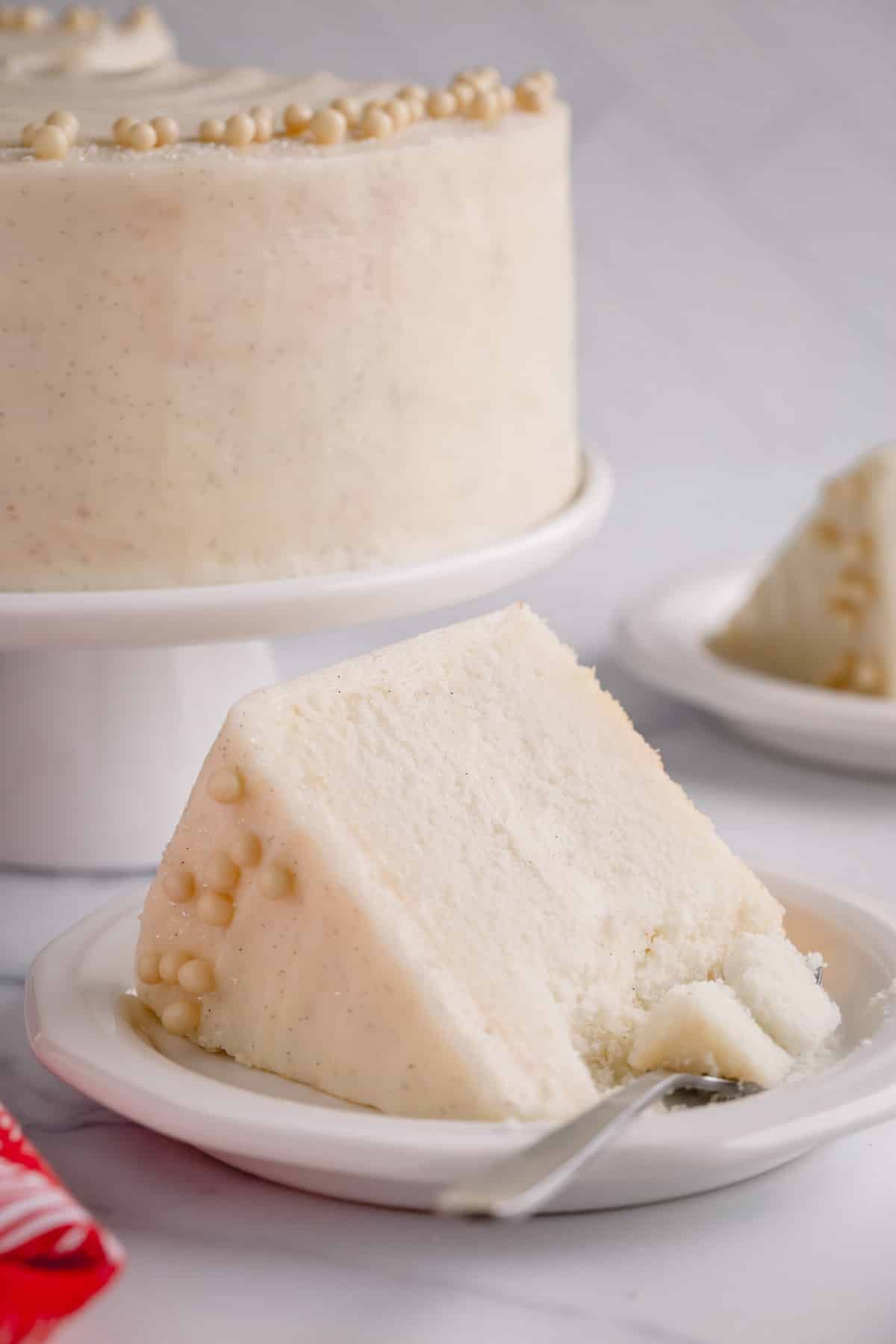 A slice of classic white cake on a plate with a fork taking a piece and the whole cake in the background.